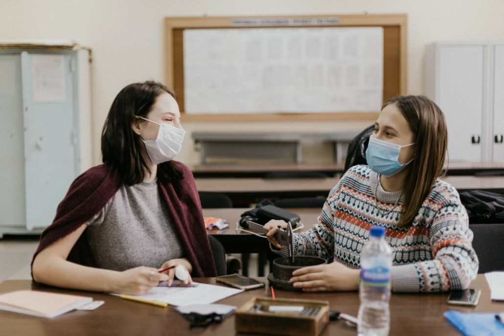 zwei junge Frauen sitzen sich in einer Schule mit Maske gegenüber, sie arbeiten an einer praktischen Aufgabe.